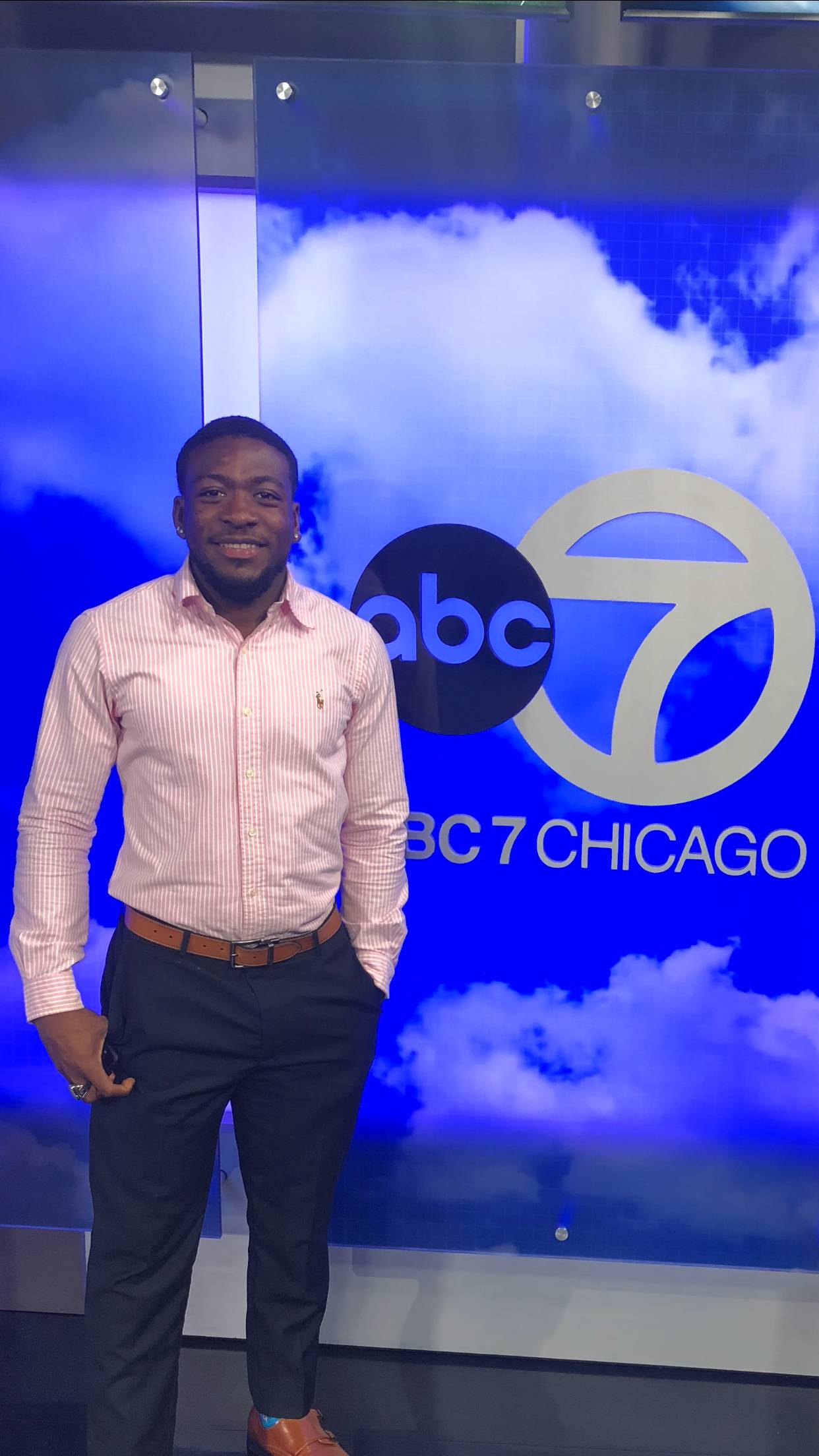 picture of a Black male standing, smiling in front of an ABC 7 Chicago logo wearing a salmon colored button down shirt and blue dress pants.
